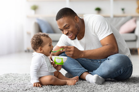 Childcare Concept. Caring Young Black Father Feeding Adorable Baby Son At Homeの写真素材