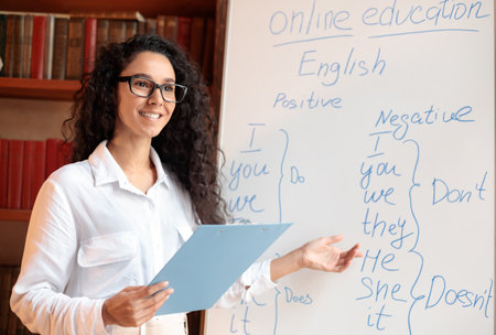 Smiling female teacher pointing at whiteboard, explaining grammar rulesの写真素材