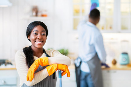 Portrait of happy African American woman with mop ready for cleanup, her husband wiping kitchen...
