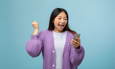Overjoyed young asian lady holding smartphone and shaking fist in joy and excitement, standing over blue backgroundの写真素材