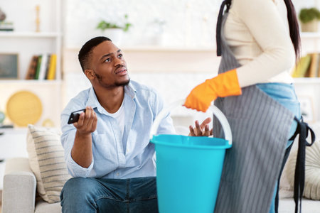 Lazy black man watching TV while his wife doing home cleanup, having fight with her, not wanting to...