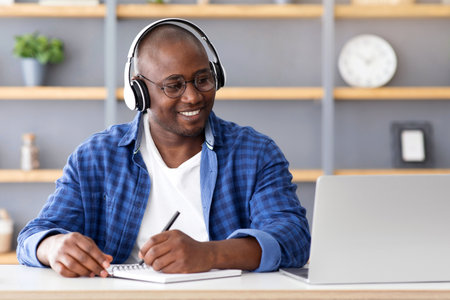 E-education. Happy black mature man using laptop and taking notes, listening online course
