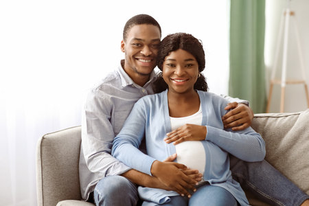 Smiling black man hugging his pregnant woman, sitting on sofaの写真素材