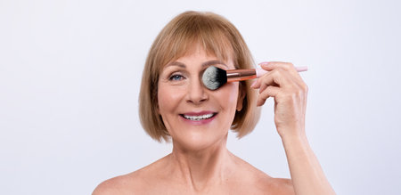 Natural makeup for aging skin. Portrait of lovely mature lady holding cosmetic brush near her eye on light backgroundの写真素材