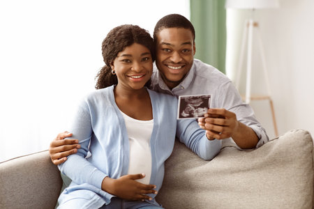 Smiling pregnant black couple holding ultrasound image, huggingの写真素材