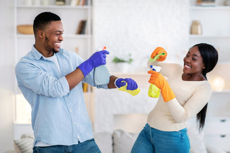 Playful black couple pretending to spray each other with detergents during house cleanup, indoorsの写真素材