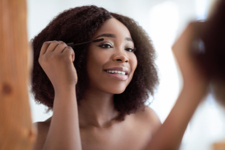 Gorgeous black woman applying mascara on her lashes in front of mirror at home