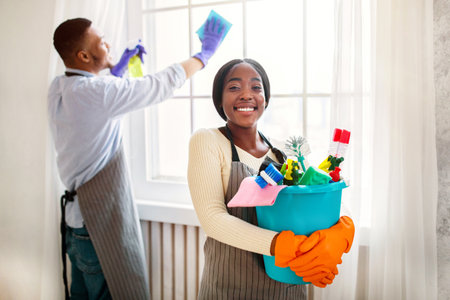 Cheerful black lady holding bucket of cleaning supplies while her husband wiping window at modern flatの写真素材