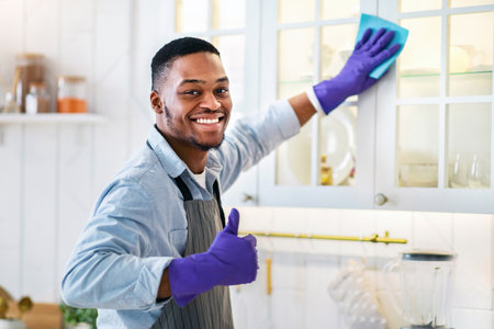Portrait of young black guy in rubber gloves tidying kitchen cabinet and showing thumb up gesture at homeの写真素材