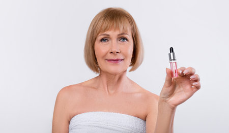 Beautiful elderly woman showing bottle with rejuvenating serum on white studio backgroundの写真素材