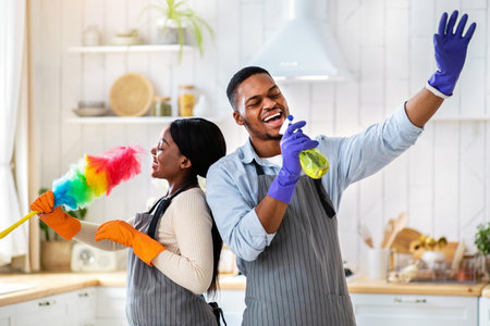 Cheerful black couple using cleaning supplies as mics, singing while tidying their home at kitchen