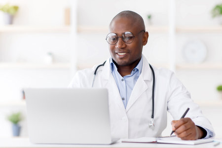 Professional help concept. Friendly black man doctor in white uniform sitting at table with laptop at his officeの写真素材