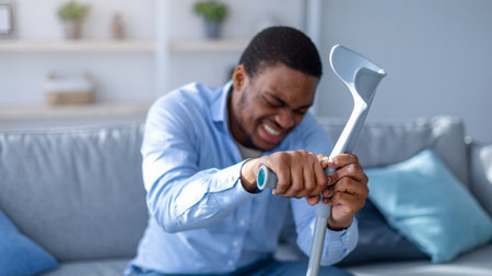 Young black man suffering from acute pain, leaning on crutch at home, selective focusの写真素材