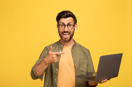 Middle aged man happily showcasing laptop against vibrant yellow backgroundの写真素材