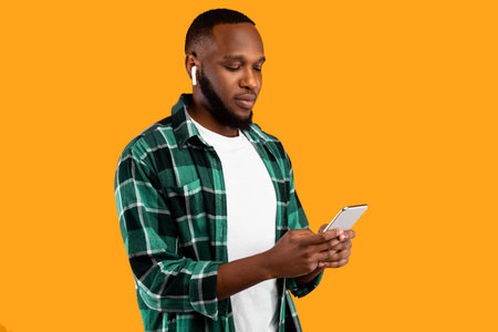 Black Millennial Man Using Cellphone Wearing Earbuds On Yellow Backgroundの写真素材