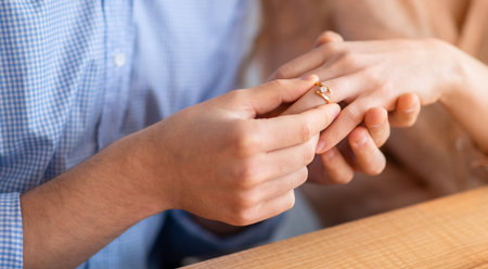 Cropped view of millennial man proposing to his beloved lady indoors, putting on diamond ring on her finger, closeupの写真素材