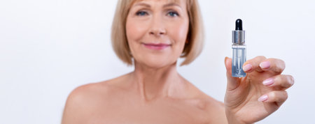 Beautiful mature woman showing bottle with rejuvenating serum on light studio background, selective...