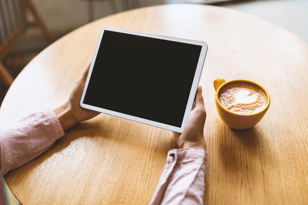 Young woman using digital tablet with black blank screen and drinking coffee, sitting at table in...