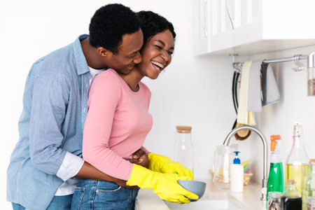 Black guy hugging his laughing girlfriend washing dishes at kitchenの写真素材