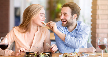 Joyful millennial guy feeding delicious salad to his beloved woman during festive dinner at cafeの写真素材