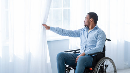 Sad with disability young man sitting on wheelchair alone at home, looking through windowの写真素材