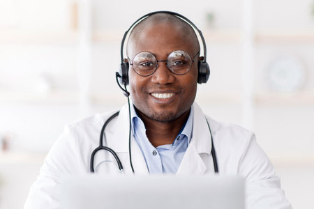 Online medical blog from home. Happy black doctor in headphones, sitting at table in interior of living roomの写真素材