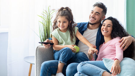 Family Weekend. Cheerful arabic parents and little daughter watching tv at homeの写真素材
