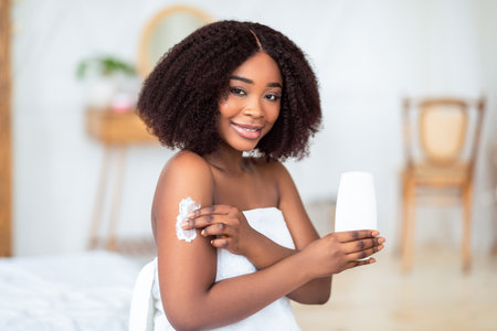 Millennial black lady applying cream, lotion or body milk after bath, making domestic beauty procedure indoorsの写真素材