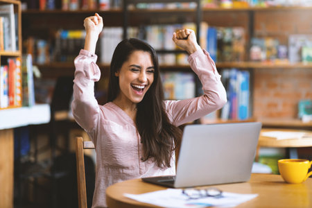 Lucky day. Emotional latin woman celebrating success in front of laptop at cafe, clenching fists and screamingの写真素材