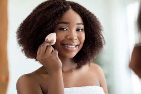 Pretty young black woman with beautiful radiant skin applying cosmetic foundation with applicator near mirror indoorsの写真素材