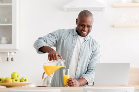 Morning routine. Happy mature black man having video chat with family on laptop computer, pouring juice in kitchenの写真素材