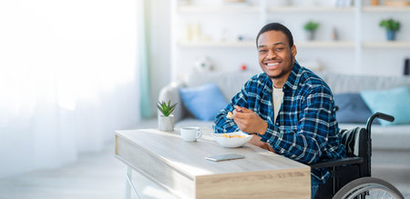 Cheerful black guy in wheelchair having breakfast, eating tasty cereal at homeの写真素材
