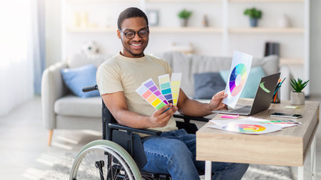 Black guy in wheelchair working as graphic designer online, holding color swatches palette, using laptop indoorの写真素材