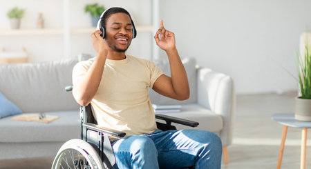 Positive black guy in wheelchair listening to music with closed eyes, using headphones at homeの写真素材