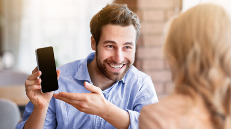 Positive millennial guy showing his girlfriend cellphone with blank screen at coffee shop, mockup for designの写真素材
