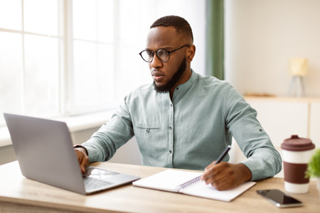 African Business Guy Working On Laptop Taking Notes At Workplaceの写真素材