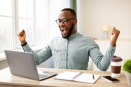 African Businessman At Laptop Shaking Fists Shouting In Joy Indoorの写真素材