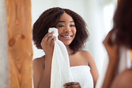 Young black woman looking at herself in mirror after shower, touching soft radiant skin on her face with towelの写真素材