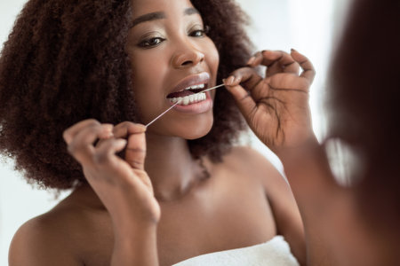 Pretty young black woman cleaning her teeth with dental floss in front of mirror at home. Oral...