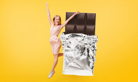 Young woman joyfully poses with giant chocolate bar on bright backgroundの写真素材