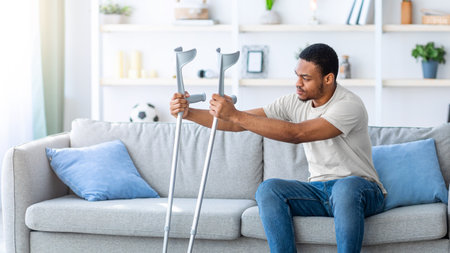 Young black guy having difficulty standing up from sofa, leaning on crutches at homeの写真素材