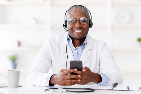 Writing patients to visit. Black man therapist in white coat with headphones using smartphone, sitting at workplaceの写真素材