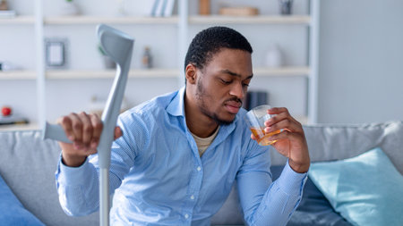 Black man with crutch suffering from pain, feeling depressed because of injury, drinking alcohol at homeの写真素材