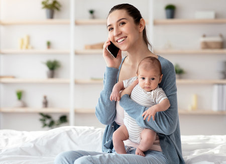 Young mothers lifestyle concept. Smiling mom talking on phone, holding baby in arms and using laptop at home.の写真素材