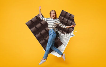 Joyful woman celebrates with chocolate bar against vibrant yellow backdropの写真素材