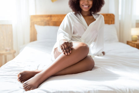 Cropped view of young black woman sitting on bed, wearing robe, touching her leg, enjoying smooth skin after depilationの写真素材