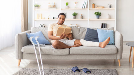 Millennial black guy with plastered leg sitting on sofa, reading book at home, pair of crutches...