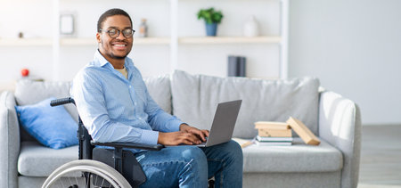 Happy with disability black man in wheelchair using laptop, working online from home, copy space
