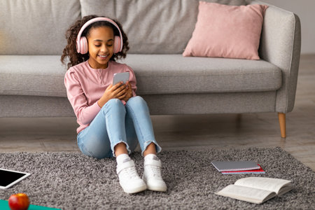 Happy african american girl listening to music in wireless headphones and chatting on her smartphoneの写真素材