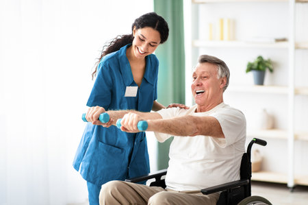 Young physiotherapist helping senior man in wheelchair do exercises with dumbbells at health centre